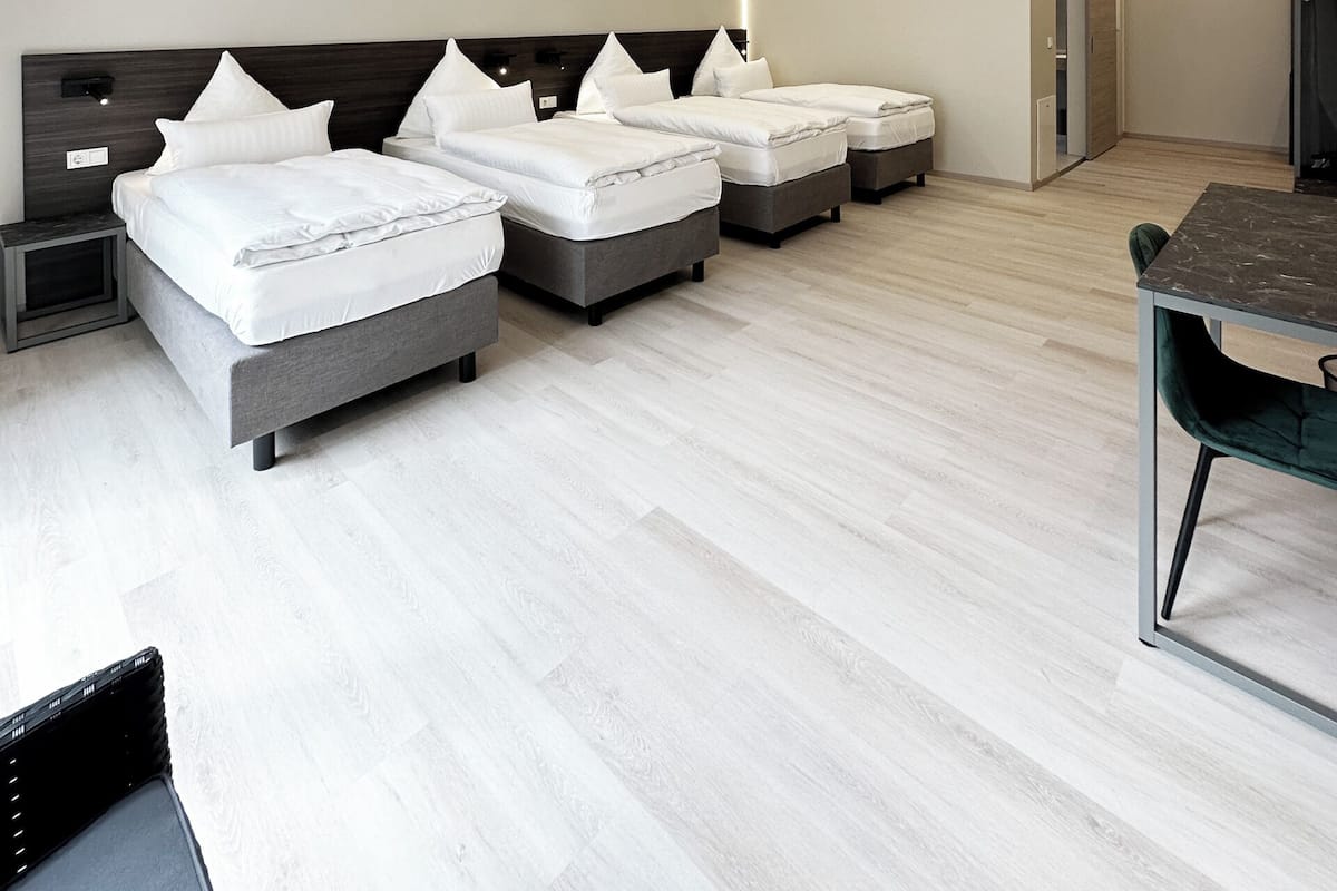 A modern hotel room with three beds, a dining table, and a small kitchen area.