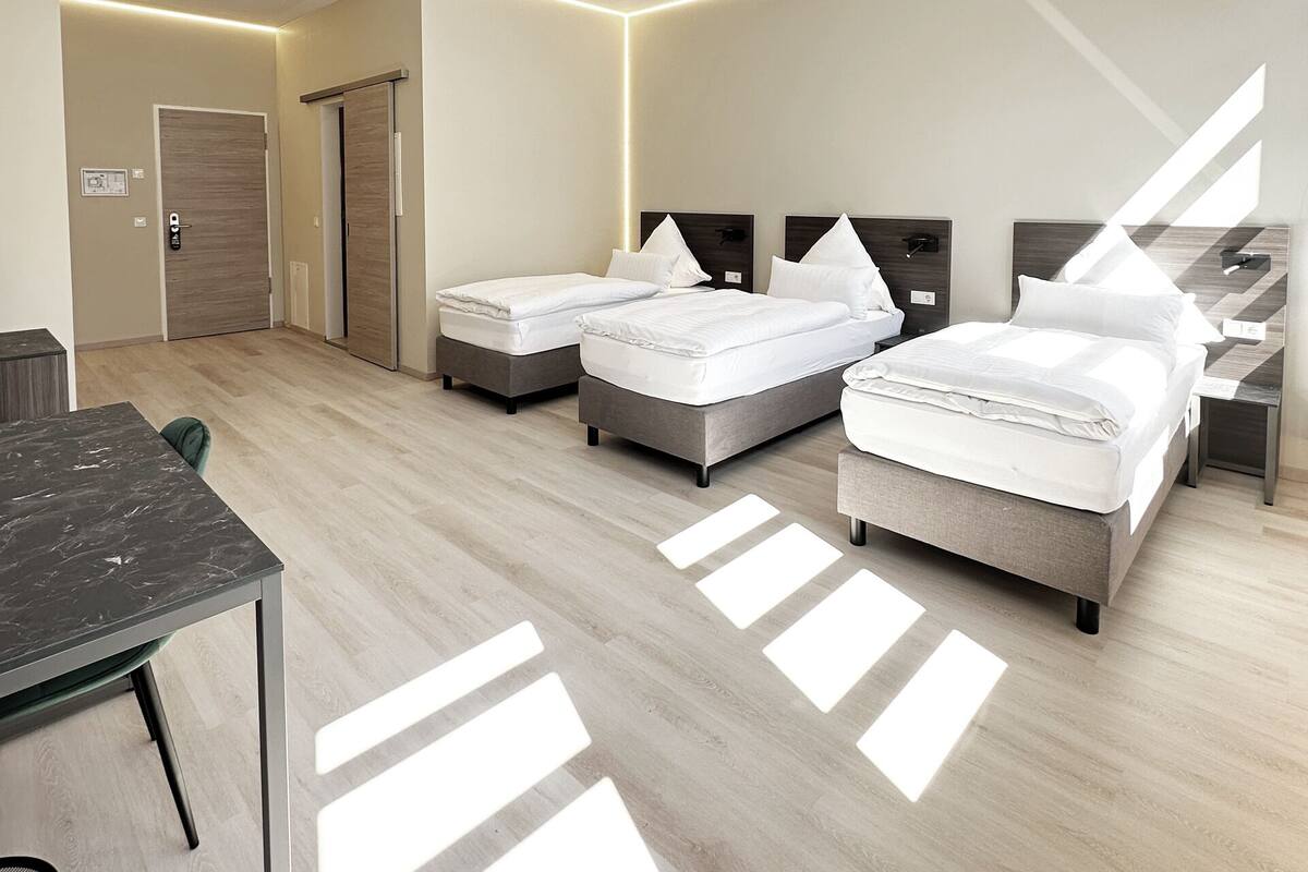 A modern hotel room with three beds, a desk, and a chair.