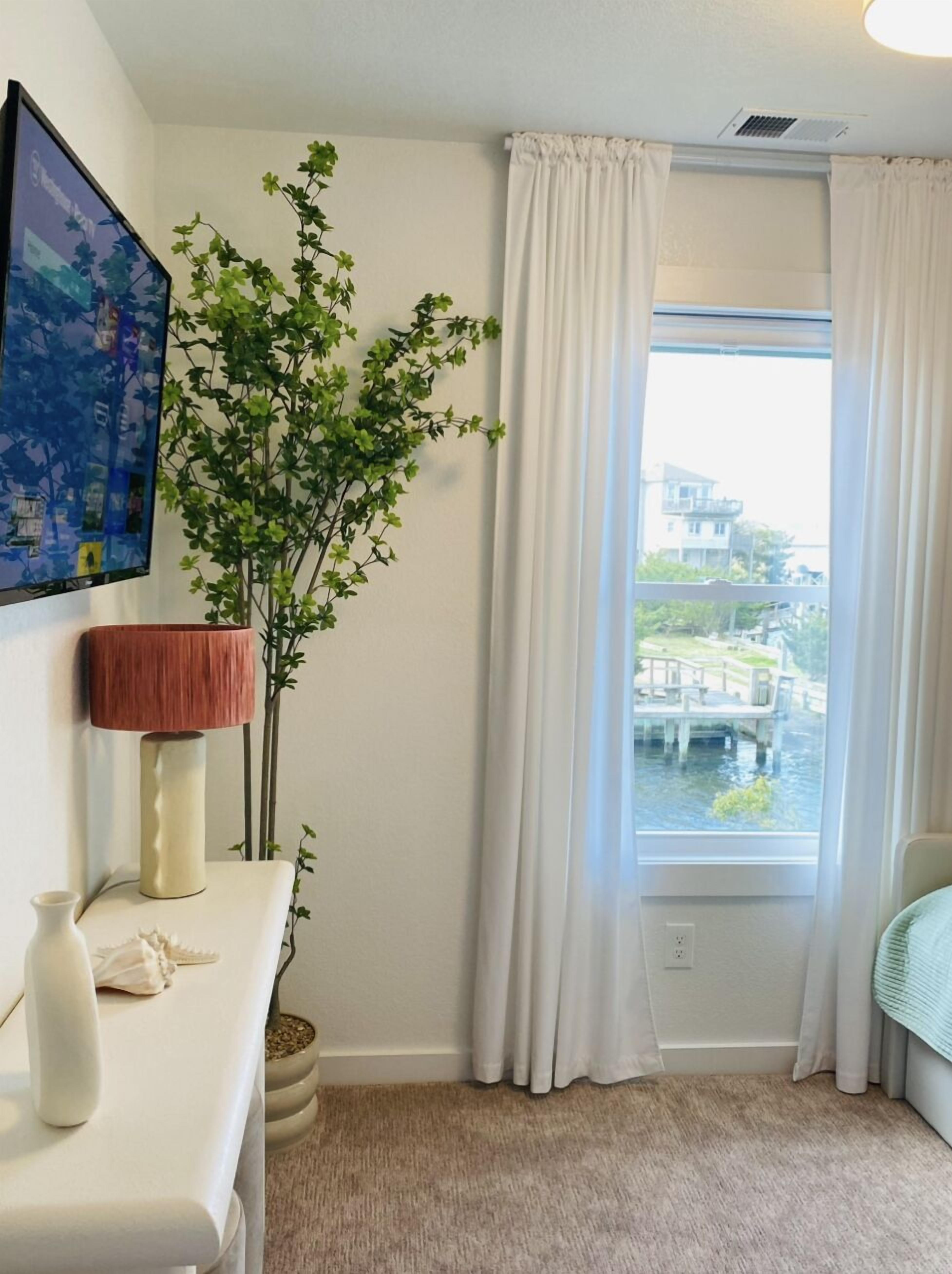 Une pièce avec une grande fenêtre, une plante en pot, une lampe et un tableau accroché au mur.