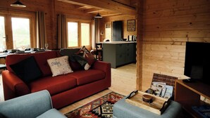 Ein gemütliches Blockhaus-Interieur mit einem roten Sofa, einem blauen Sessel, einer Küchenzeile und einem Esstisch für vier Personen.