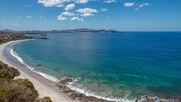 Un paisaje costero con mar azul cristalino, playa arenosa y montañas a lo lejos.