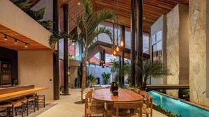 A modern indoor pool area with a wooden table and chairs, surrounded by stone walls and tropical plants.