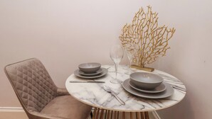 A round dining table with marble top, surrounded by a tufted chair, a gold vase, and a simple table setting with plates and cutlery. - The Salisbury Apartment 103 (Scarborough)
