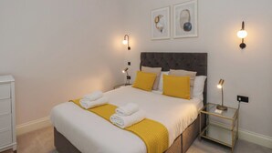 A neatly made bed with white and yellow linens, a tufted headboard, and two wall-mounted lamps. - The Salisbury Apartment 103 (Scarborough)