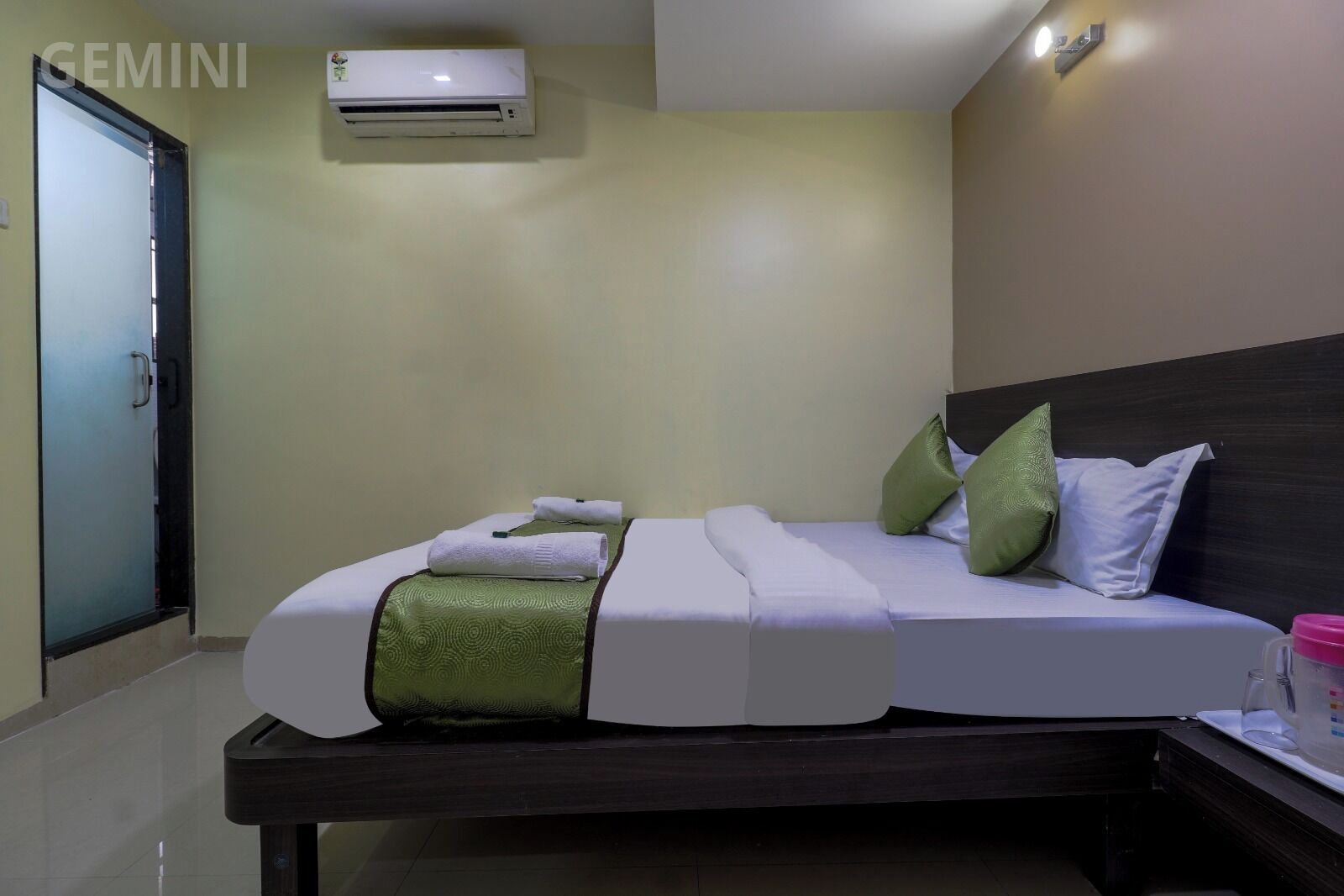 Une chambre d’hôtel avec un lit, une table de chevet, un plaid vert et un appareil de climatisation fixé au mur.