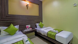 A hotel room with two beds, each with green and white bedding, a nightstand with a phone and a small lamp, and a wall-mounted light fixture above the beds. - HOTEL GEMINI (Mumbai)