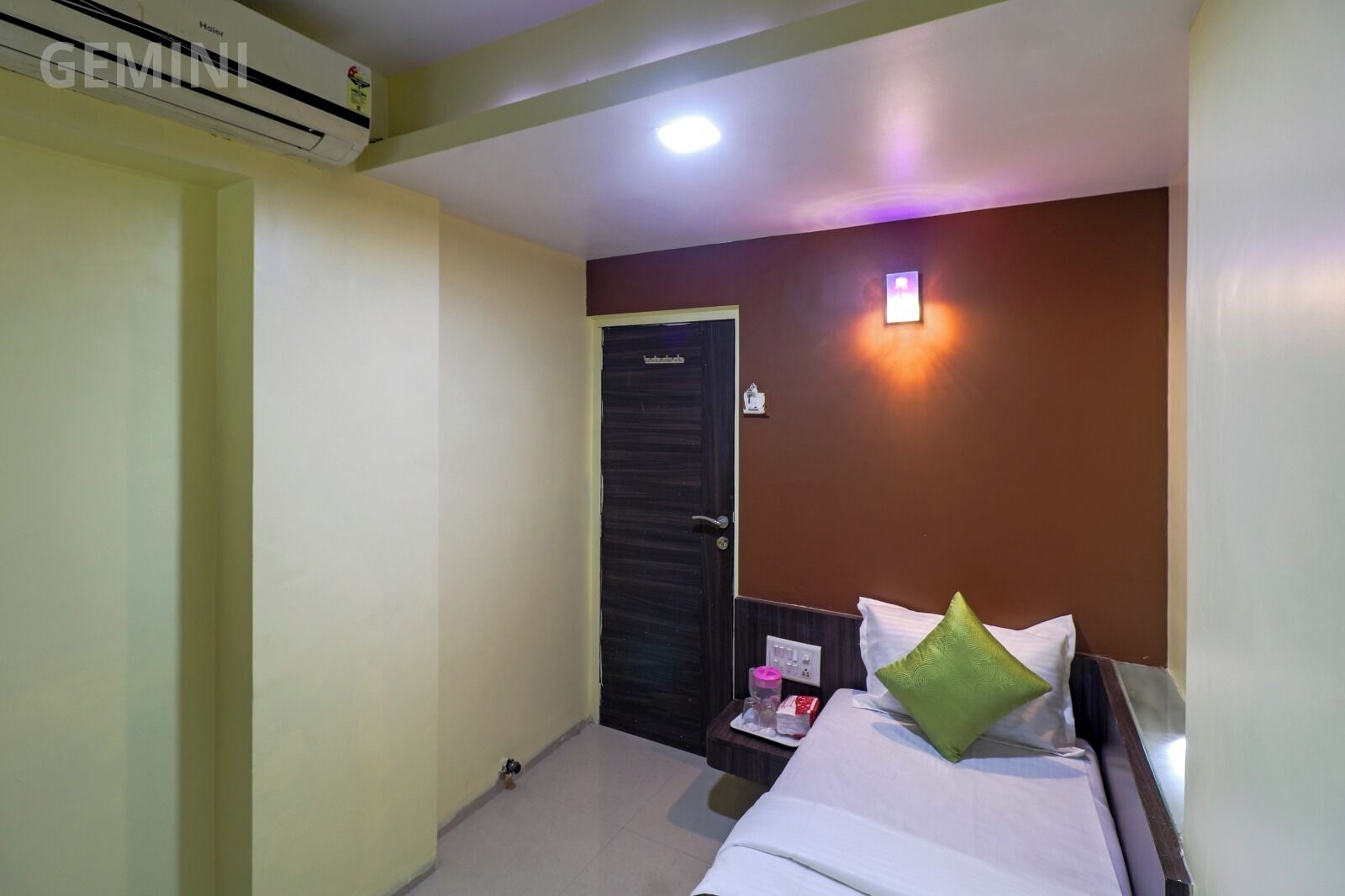 A hotel room with a bed, a nightstand, a wall-mounted light, and a door.
