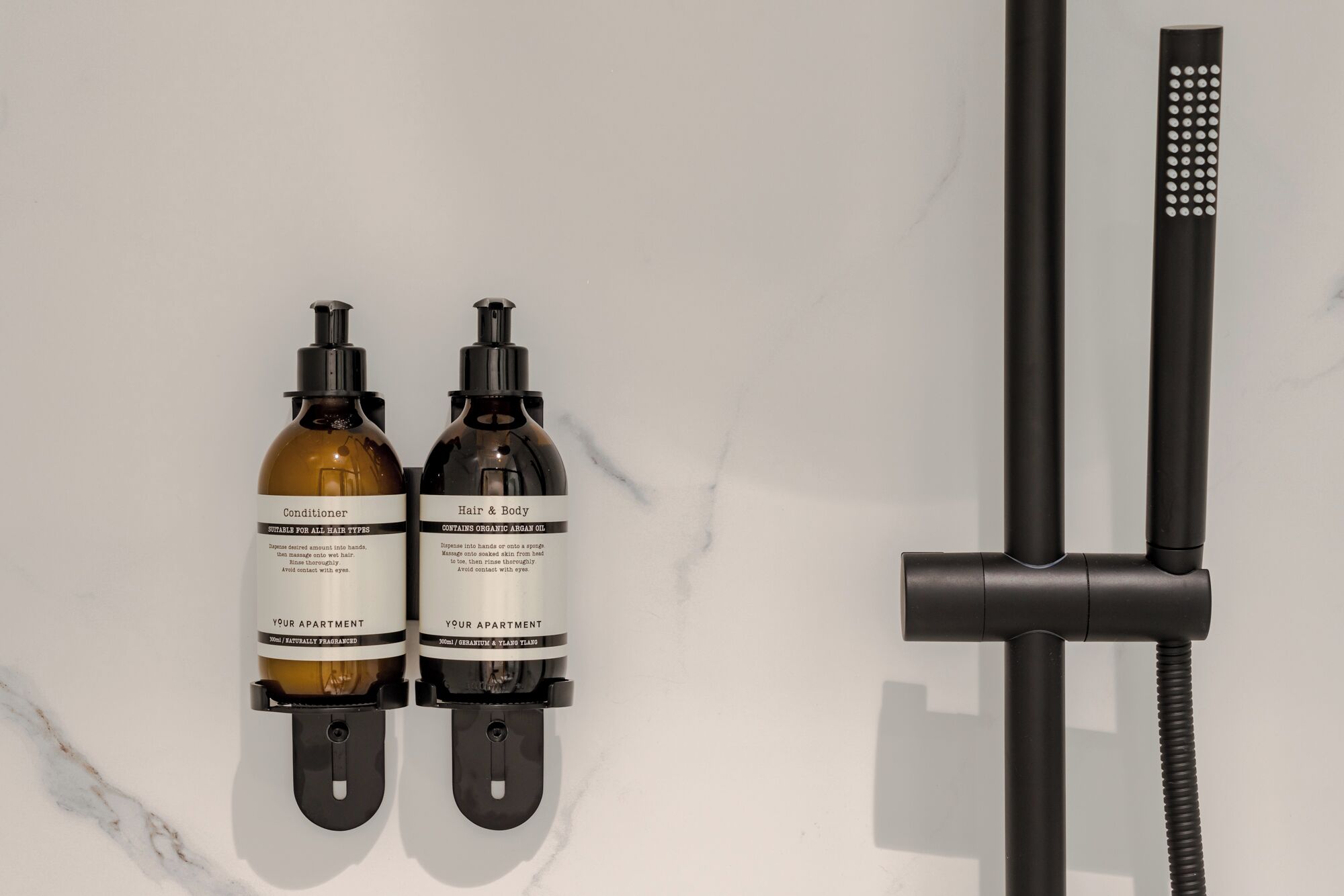 Two bottles of conditioner and a showerhead on a white marble wall.