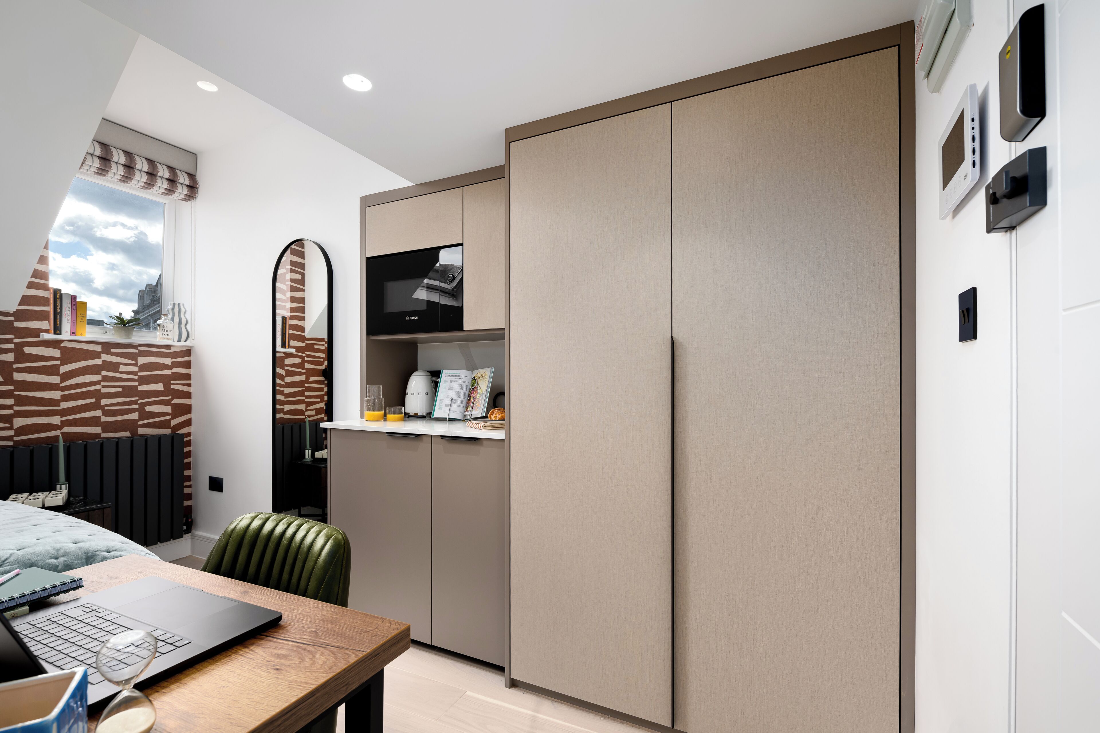 A modern bedroom with a bed, a desk with a laptop, a green chair, and a wardrobe.