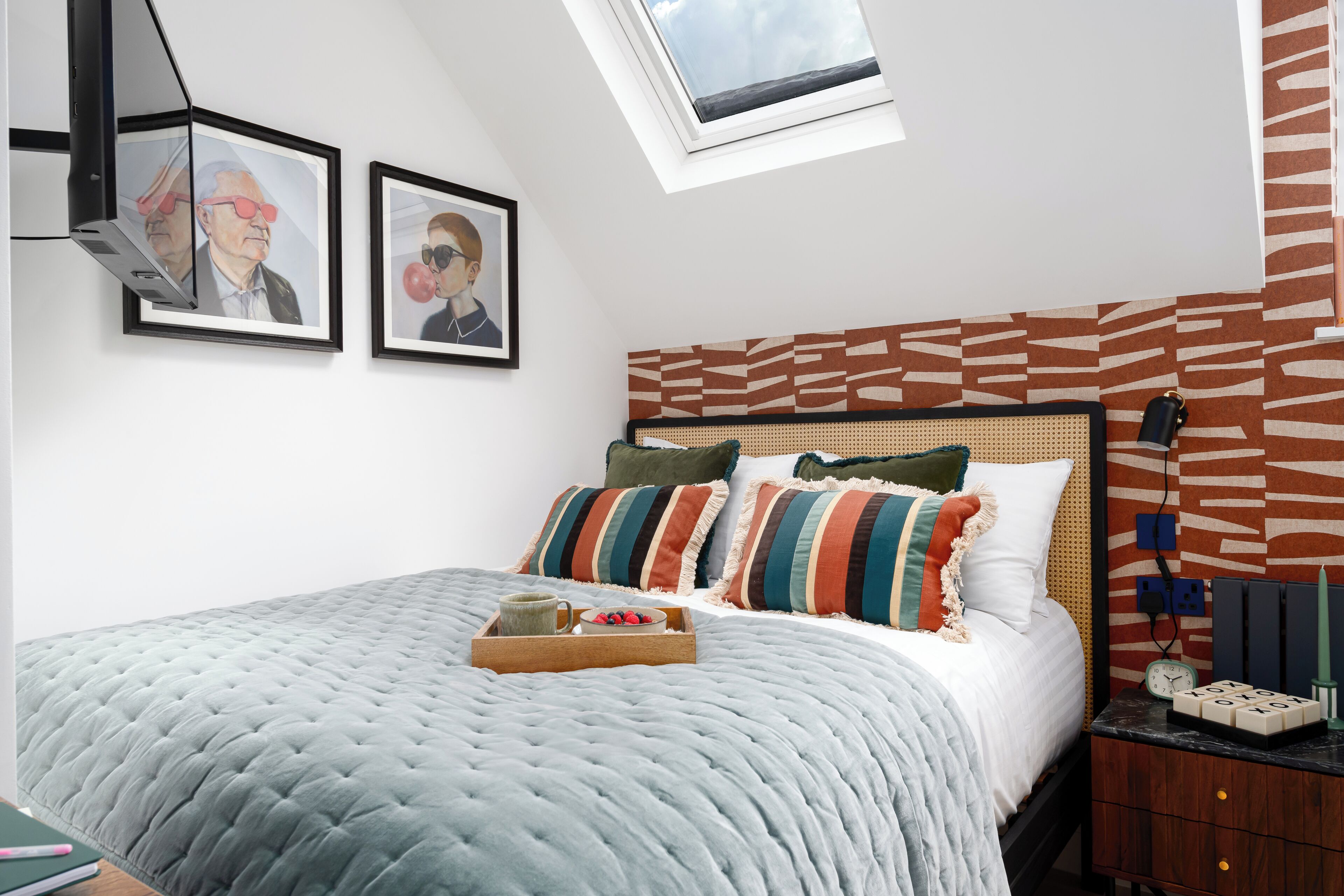 A bedroom with a bed, a bedside table, a tray with a mug and snacks, a clock, and framed pictures on the wall.