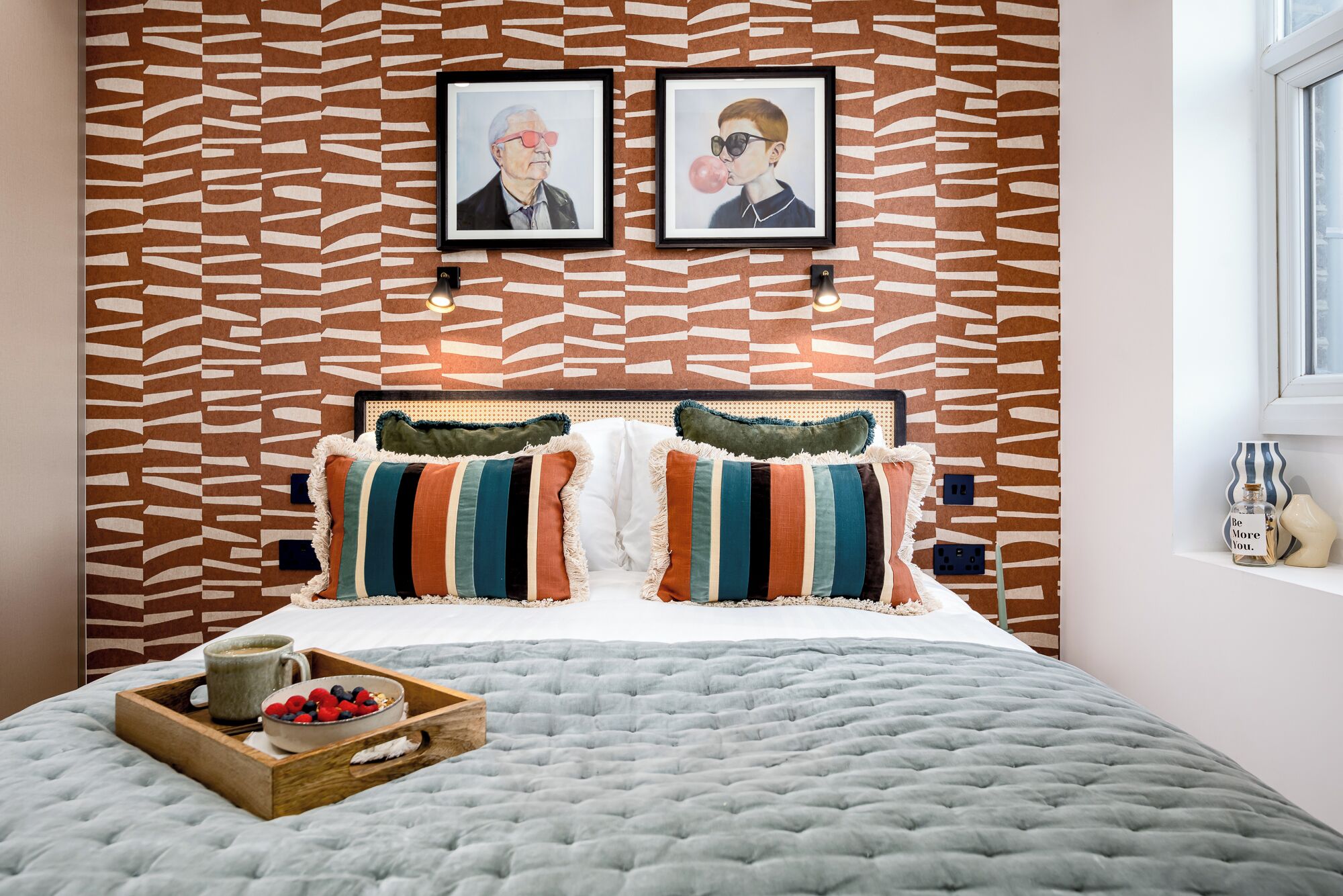 Un dormitorio con pared estampada, cama con almohadas a rayas, mesita de noche con lámpara y ventana.