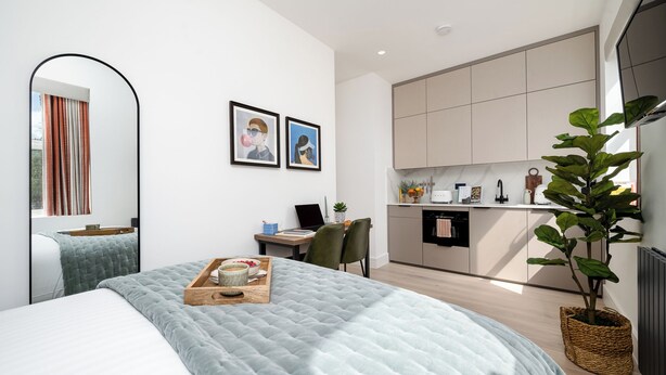 A modern bedroom with a large bed, a desk with a laptop, and a view of a kitchen and living area. - Your Apartment - Camden  (London)