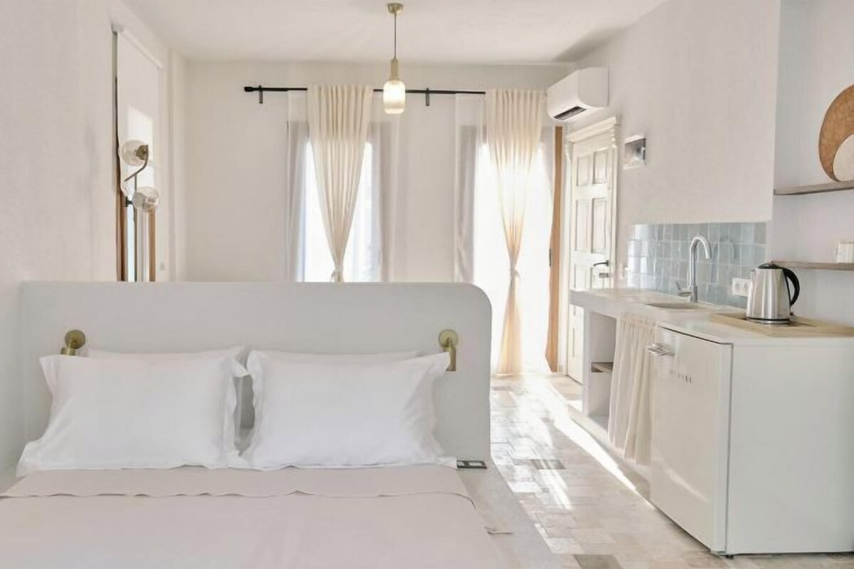 Une chambre à coucher comprenant un lit, une table de chevet, une commode, une fenêtre avec des rideaux et une vue sur un coin cuisine.