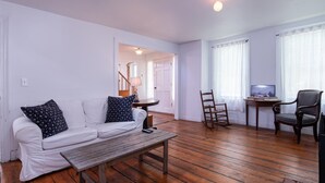 Una sala de estar con un sofá blanco, un banco de madera, un televisor sobre una mesita y una silla.