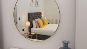 A bedroom with a round mirror, a bed with white bedding, a grey tufted headboard, and two yellow pillows.