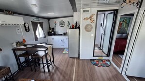 A compact kitchen with a refrigerator, a dining table with chairs, and a small island. There is a clock on the wall and a window with curtains.