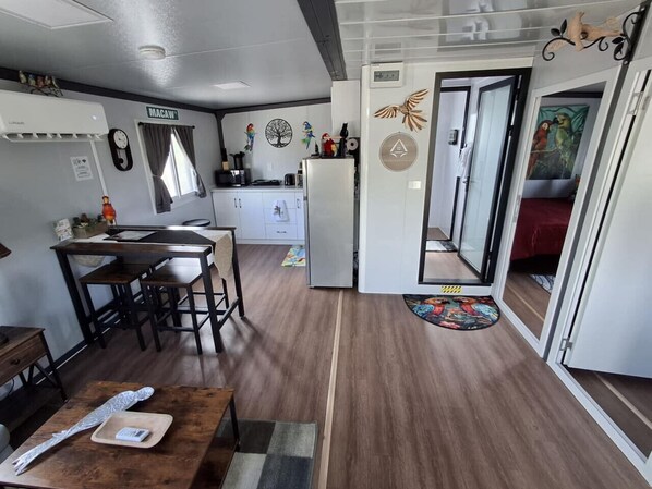 A compact kitchen with a refrigerator, a dining table with chairs, and a small island. There is a clock on the wall and a window with curtains. - The Hideaway at Arrowhead (#3) (Sandia)