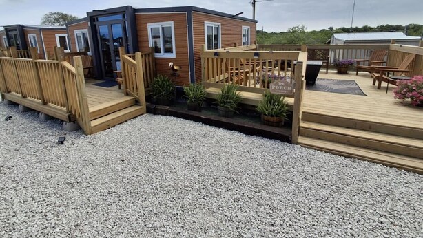 A modern mobile home with a wooden deck, a small garden area, and a sign indicating a 'Pool Area'.
