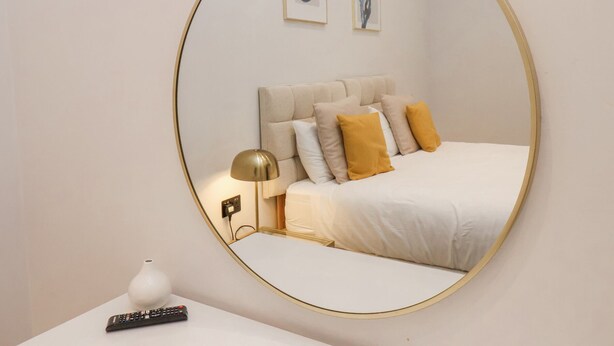 A bedroom with a large oval mirror, a bed with multiple pillows, a bedside table with a lamp, and a remote control. - The Salisbury Apartment 106 (Scarborough)