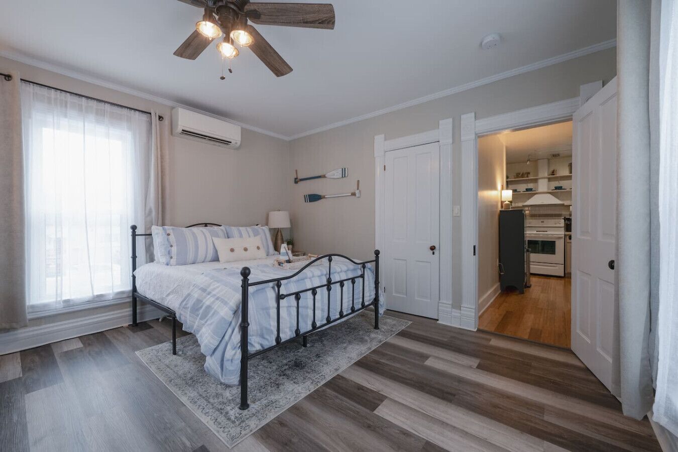 A bedroom with a bed, a ceiling fan, an air conditioning unit, and a view into the kitchen.