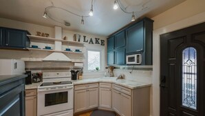 Cocina con armarios blancos, estufa y microondas. Hay estantes con vajilla y una ventana.