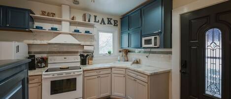 A kitchen with white cabinets, a stove, and a microwave. There are shelves with dishes and a window.