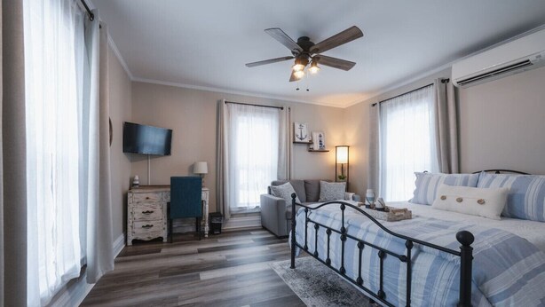 Un dormitorio con cama, escritorio, silla, televisor, ventilador de techo y ventana.