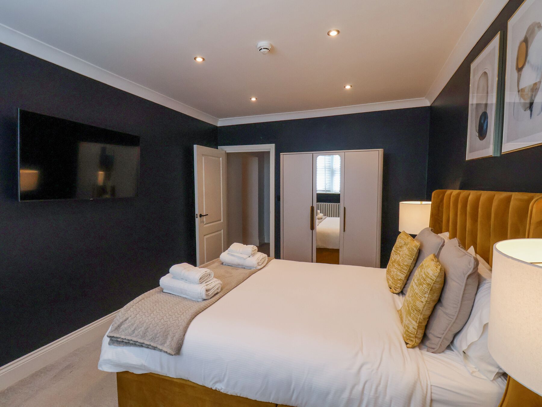 A modern bedroom with a large bed, a flat-screen TV mounted on the wall, and a wardrobe with a mirror.