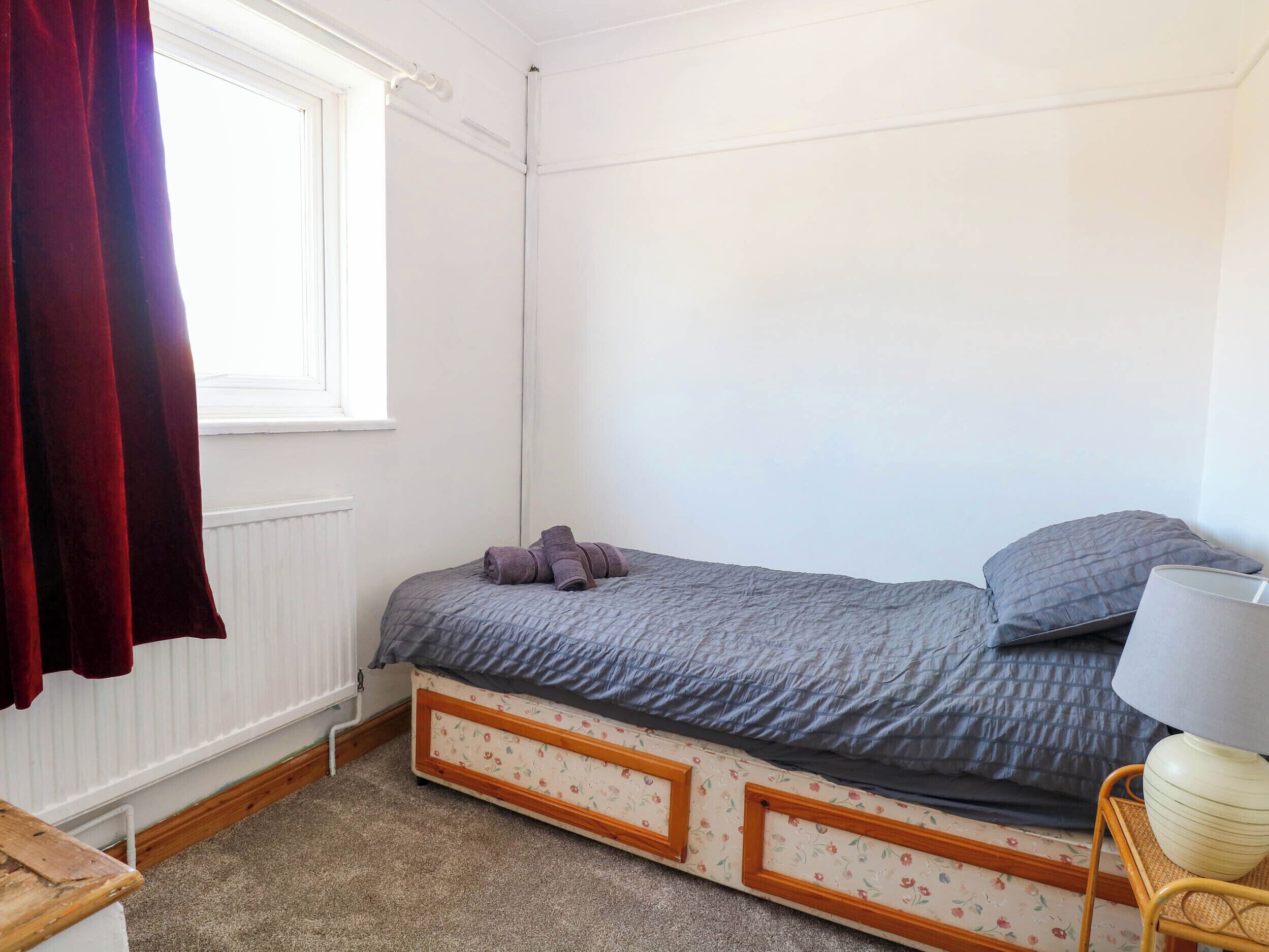 A small bedroom with a single bed, a window with curtains, a radiator, and a lamp on a side table.