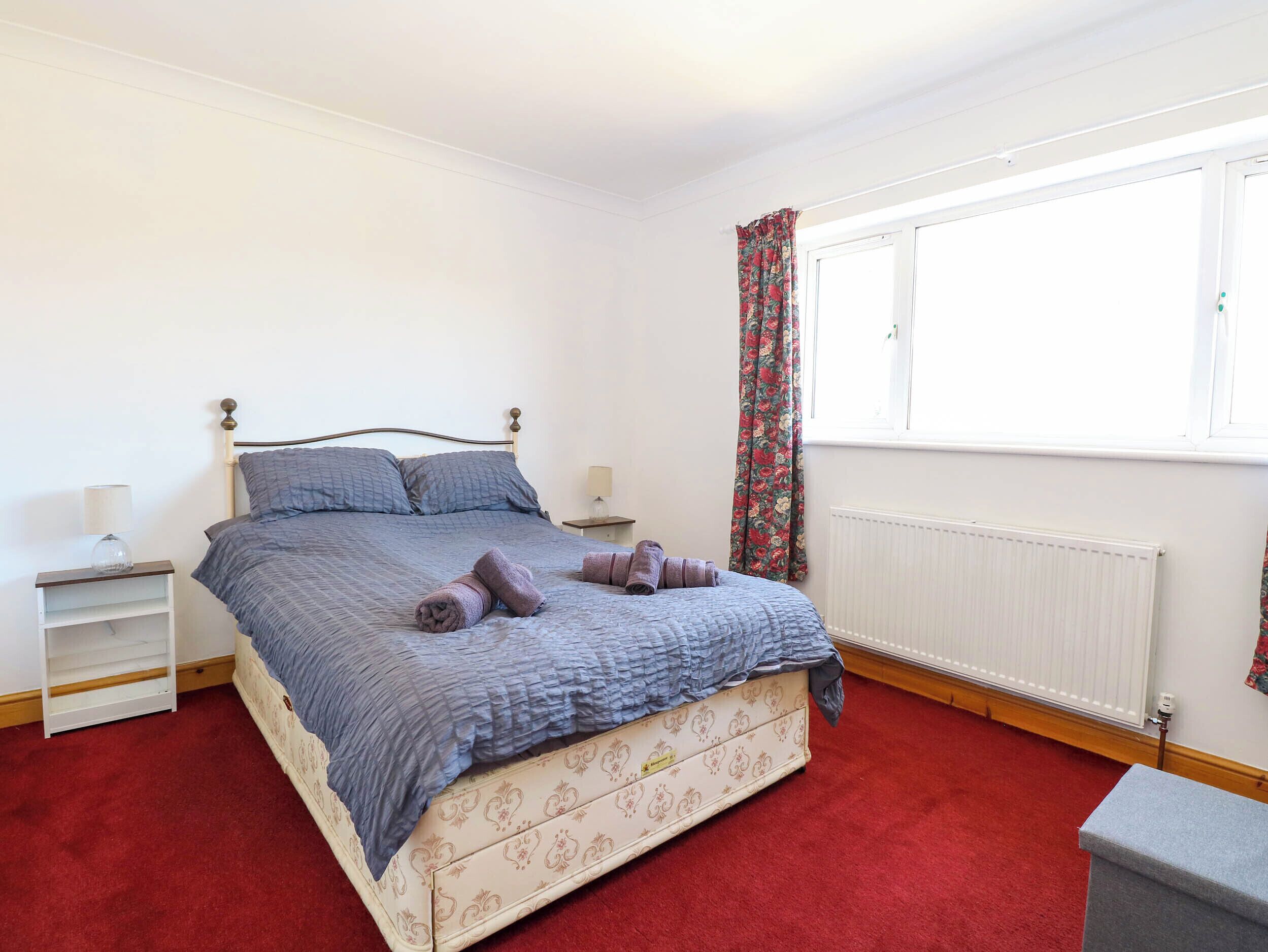 A bedroom with a bed, a nightstand, a lamp, a window with curtains, and a radiator.