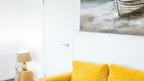 Een gele bank, een houten bijzettafeltje, een lamp en een schilderij van een boot op het strand.