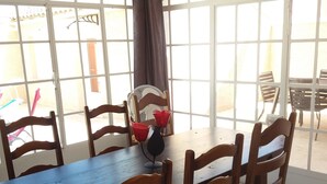 Un coin repas avec une table et des chaises en bois, de grandes fenêtres avec des rideaux et un ventilateur.