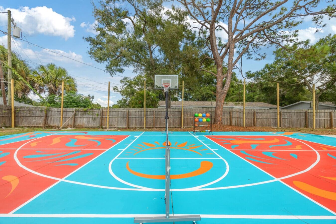 An outdoor basketball court with a wooden fence, trees, and a clear sky.