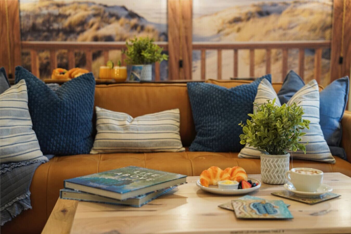 Un coin salon accueillant avec un canapé en cuir, des coussins à rayures, une petite table avec des livres, une assiette de croissants, une tasse et une plante en pot.