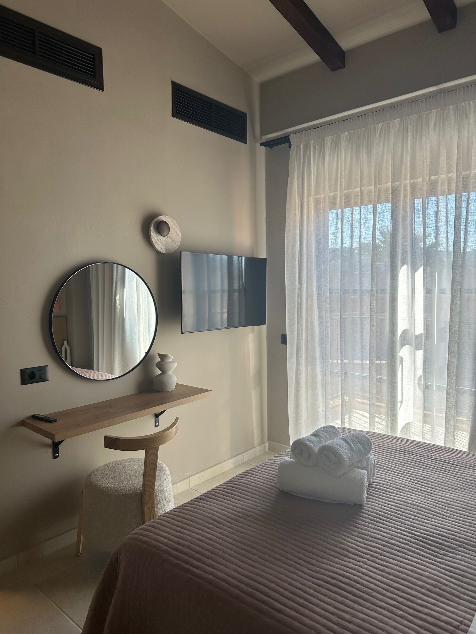 A bedroom with a bed, a bedside table, a mirror, a TV, and a window with curtains.