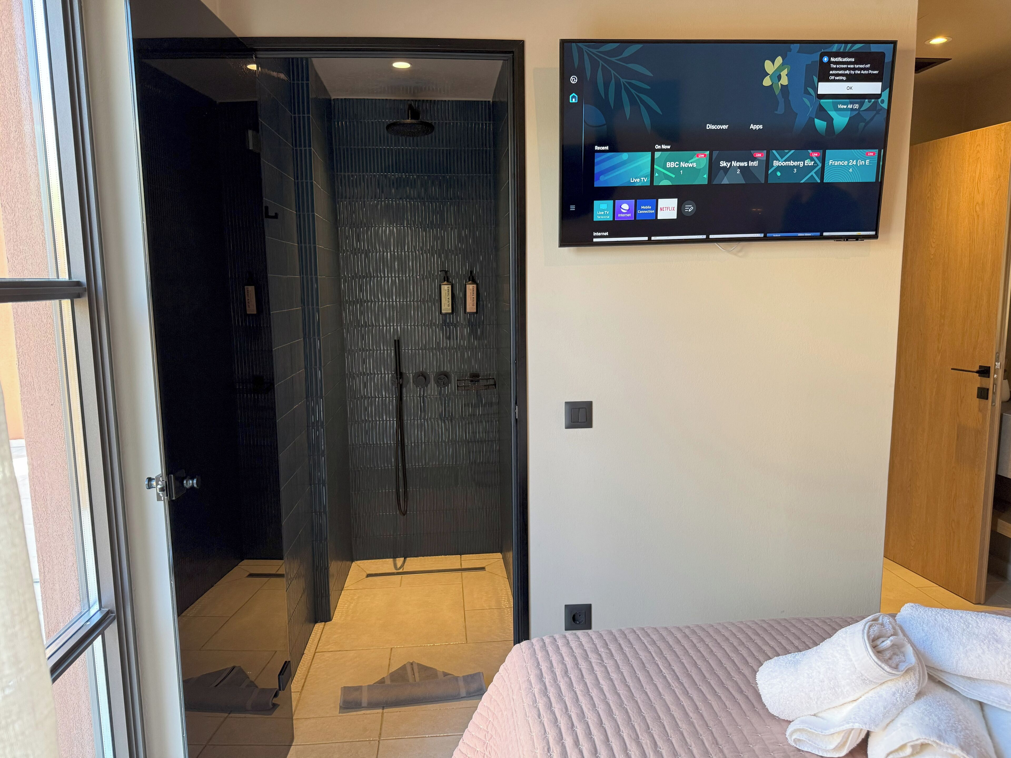 Ein Hotelzimmer mit Glastür zum Duschbereich, einem an der Wand befestigten Flachbildfernseher, der einen Nachrichtensender zeigt, und einem Bett mit ordentlich gefalteten Handtüchern.