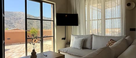 A modern living room with a large window, a flat-screen TV, and a sofa with cushions.