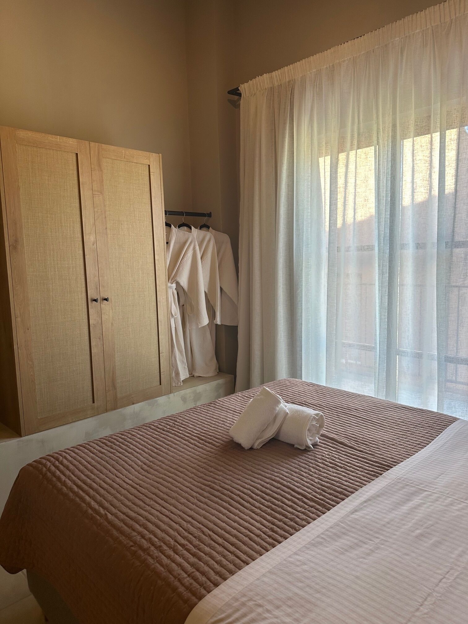 A bedroom with a large bed, a wooden wardrobe, and a window with sheer curtains.