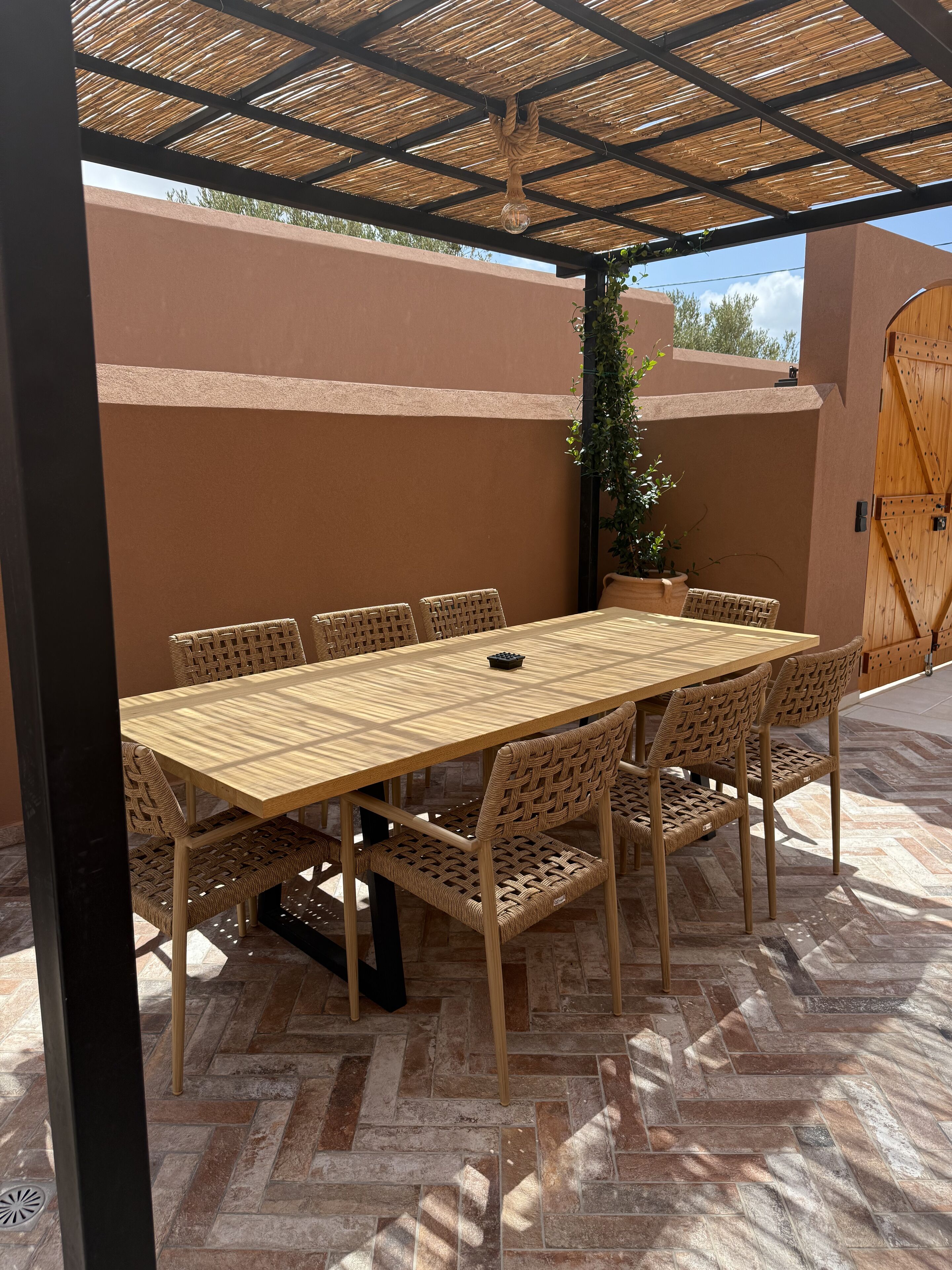 Uma área de refeições ao ar livre coberta, com mesa de madeira e cadeiras de fibra, chão de azulejos e um portão de madeira ao fundo.
