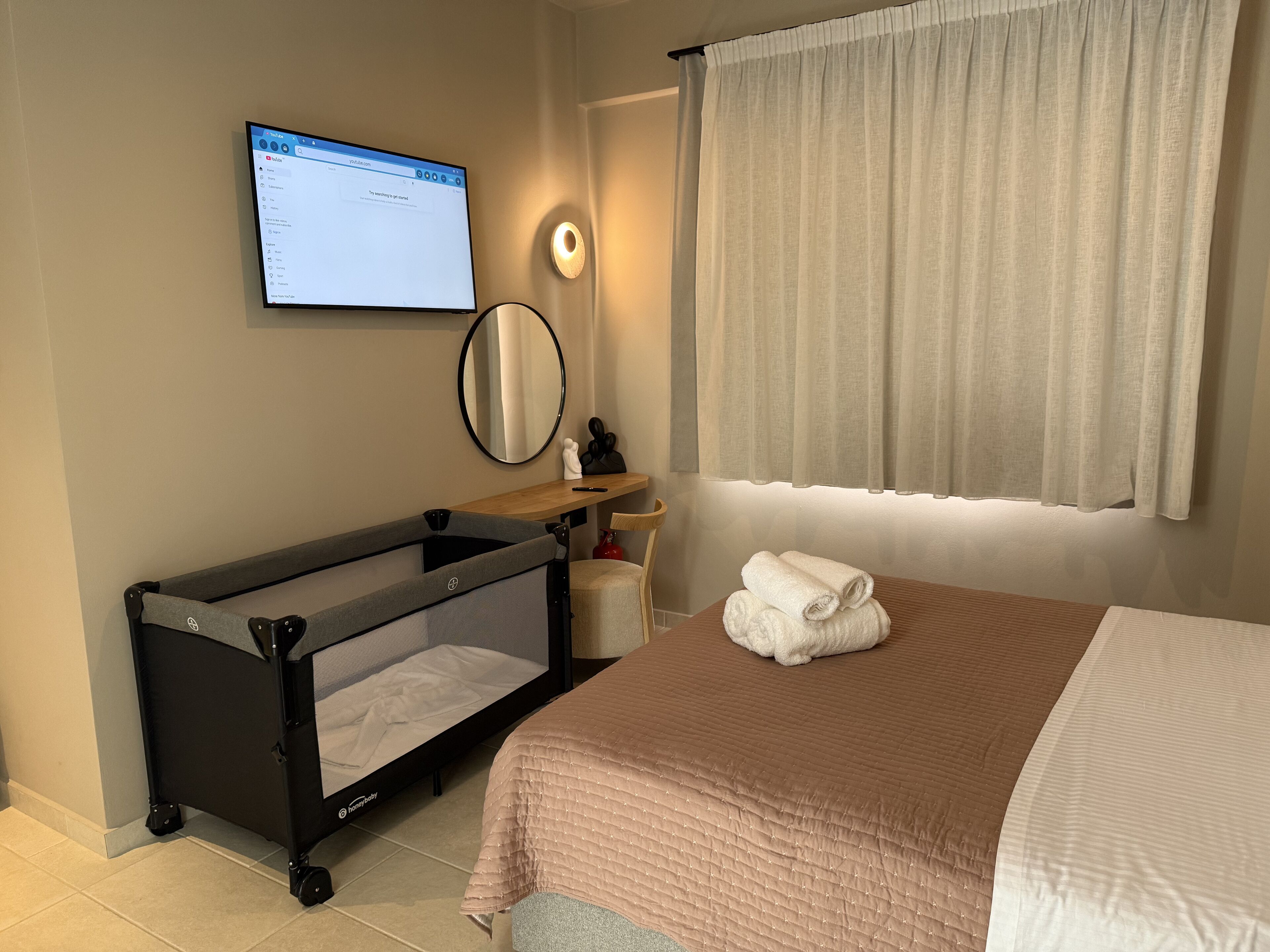 Une chambre d’hôtel avec un berceau, un lit recouvert d’une couverture rose, un téléviseur fixé au mur, un miroir et une fenêtre avec des rideaux.