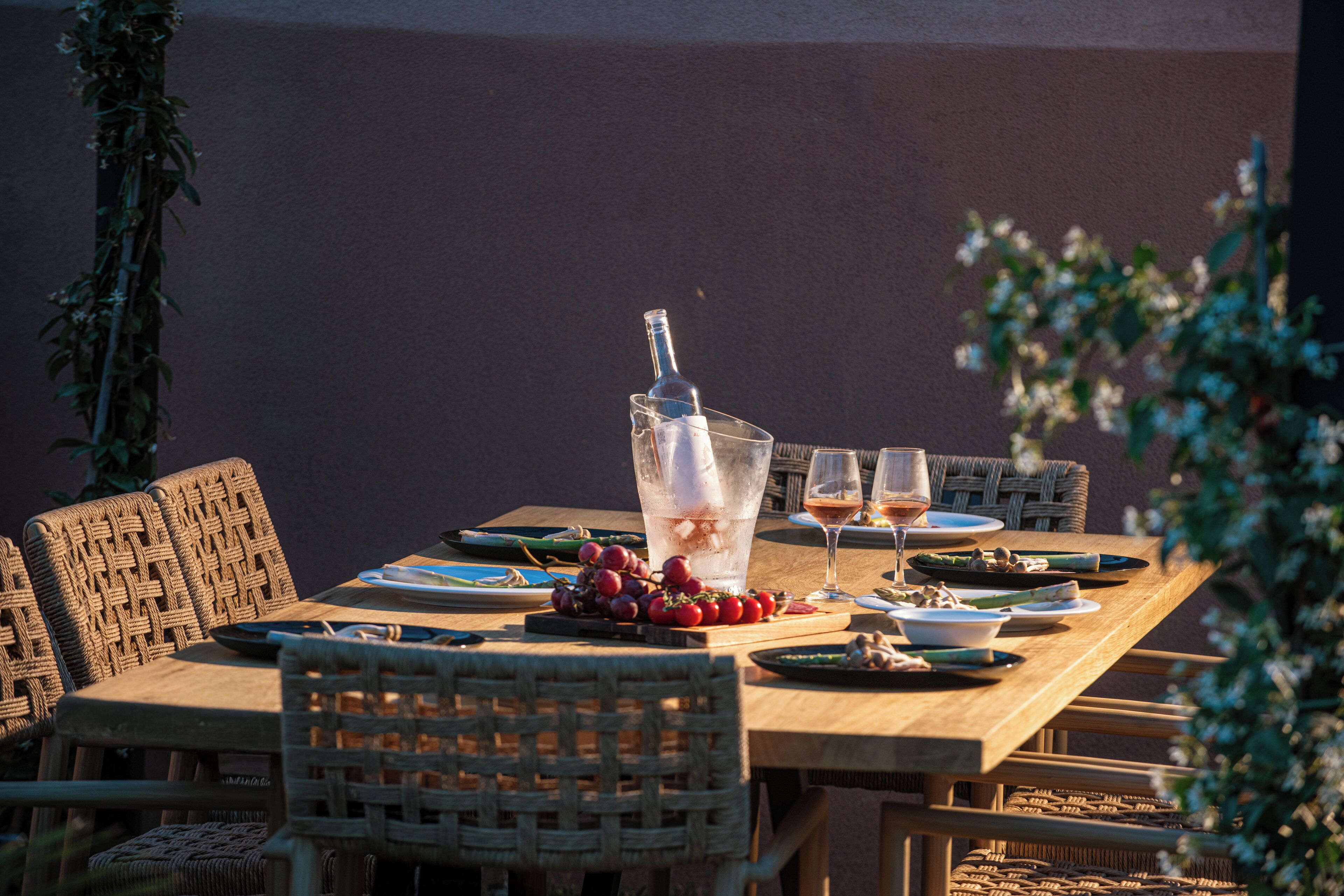 A wooden dining table set with wicker chairs, a bottle of champagne, glasses, and a bowl of grapes.