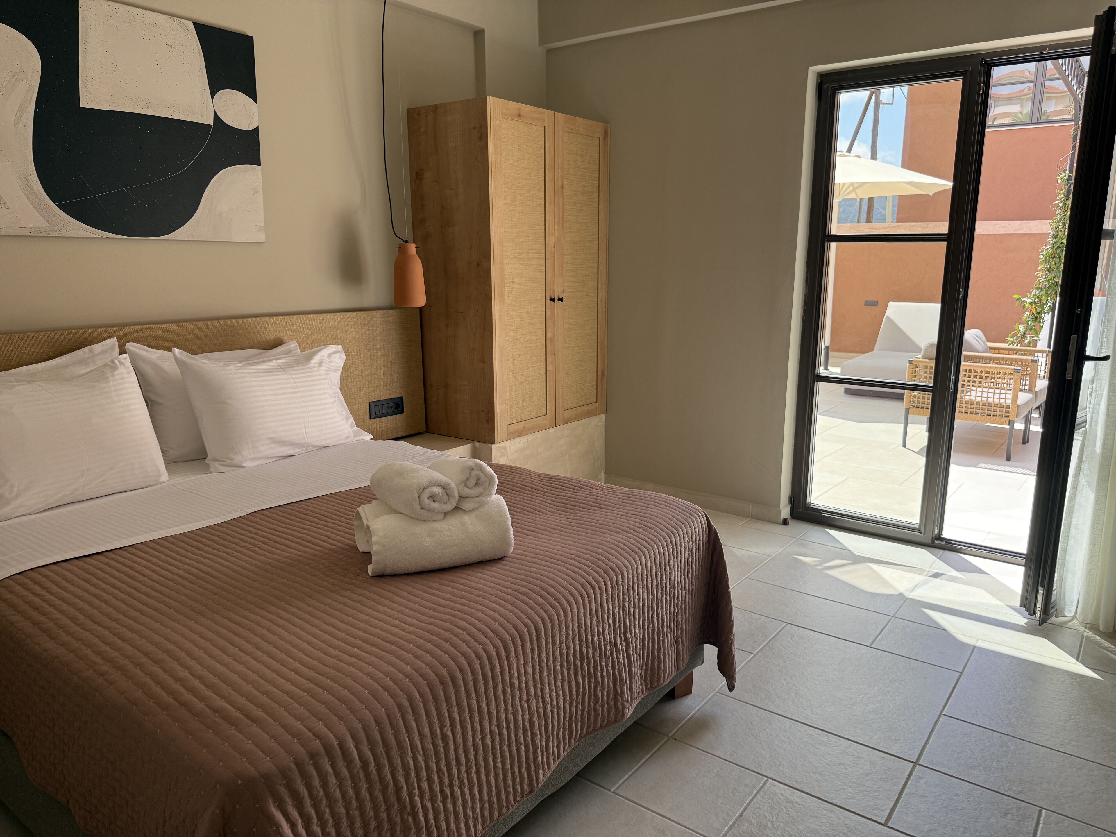 Ett hotellrum med en stor säng, en garderob i trä och utsikt över uteserveringen genom en glasdörr.