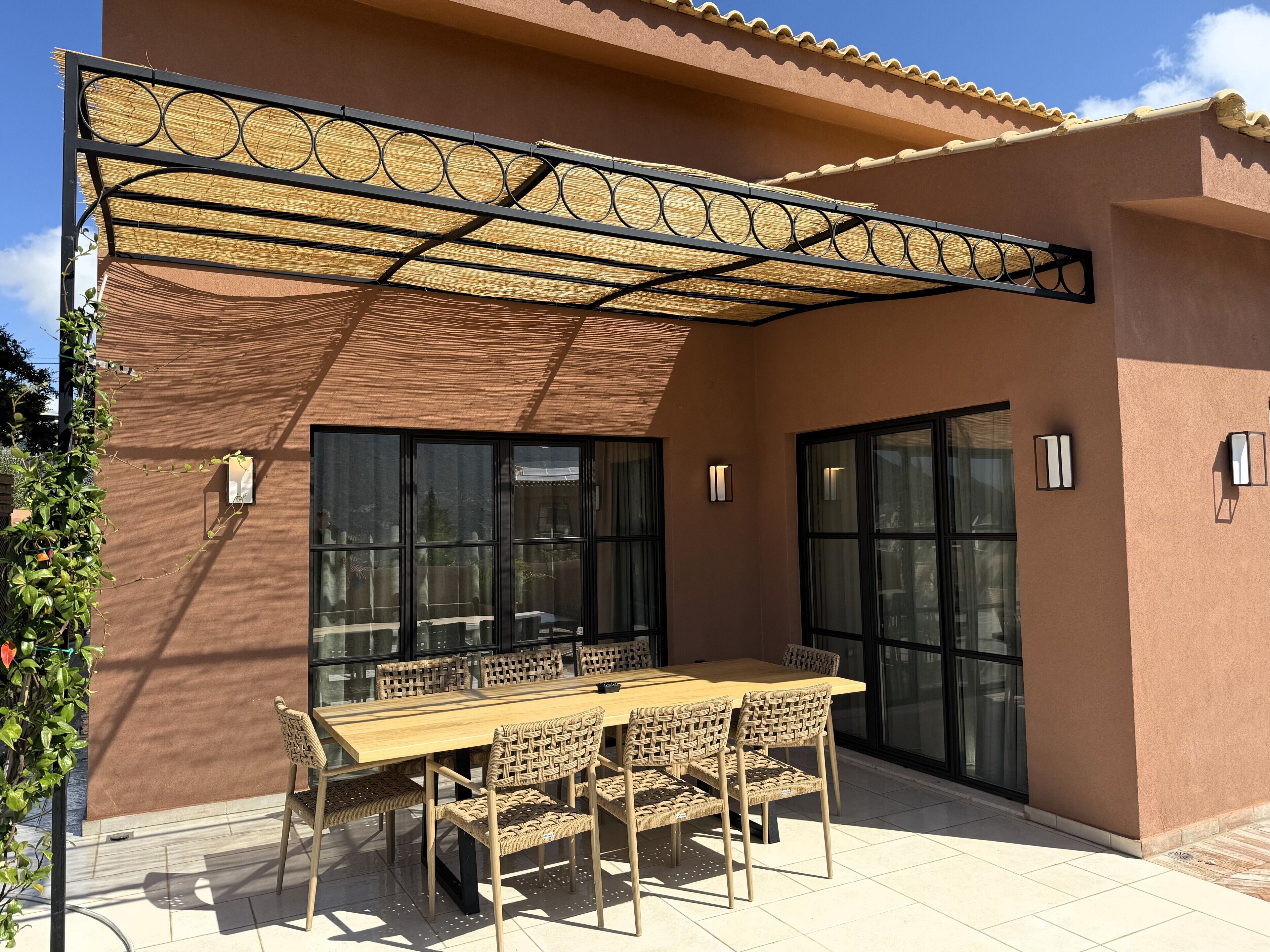 Patio esterno con tavolo in legno e sedie in vimini, zona coperta con tenda in fibra intrecciata e ampie finestre.