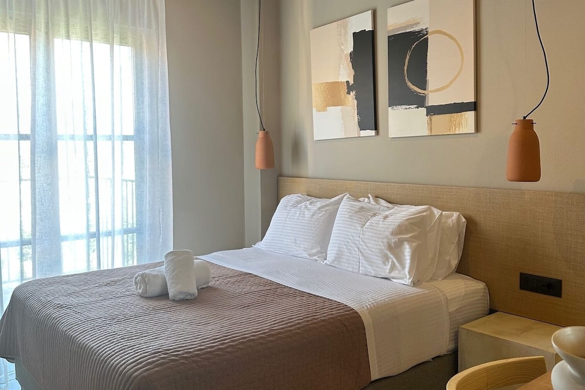 A neatly made bed with white linens, a brown throw blanket, and a white towel on the side. There are two wall-mounted artworks above the bed, a wooden headboard, and a small bedside table with a vase.