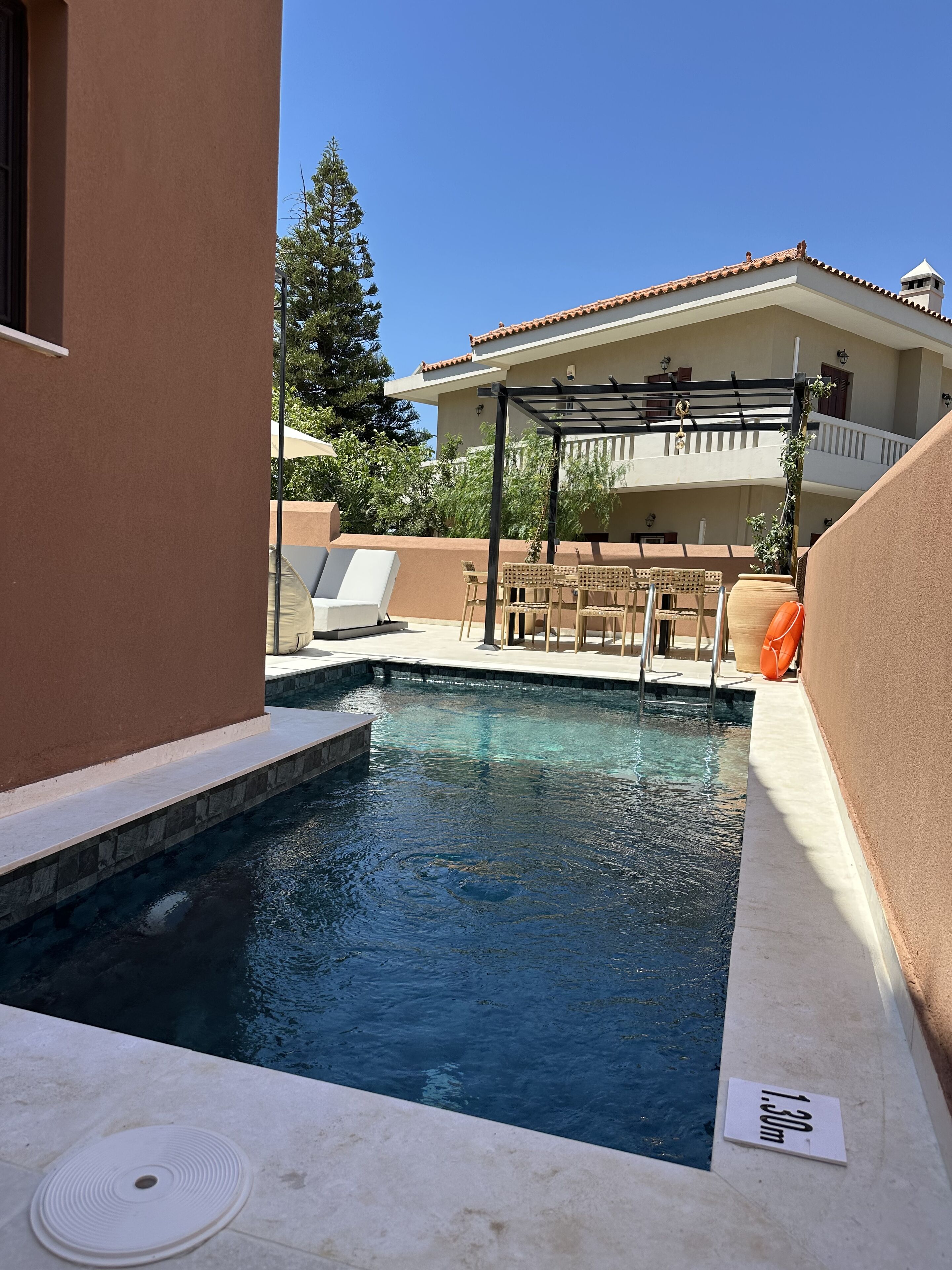 Una piscina con vista a aguas cristalinas, una pérgola con asientos y un edificio de dos pisos al fondo.