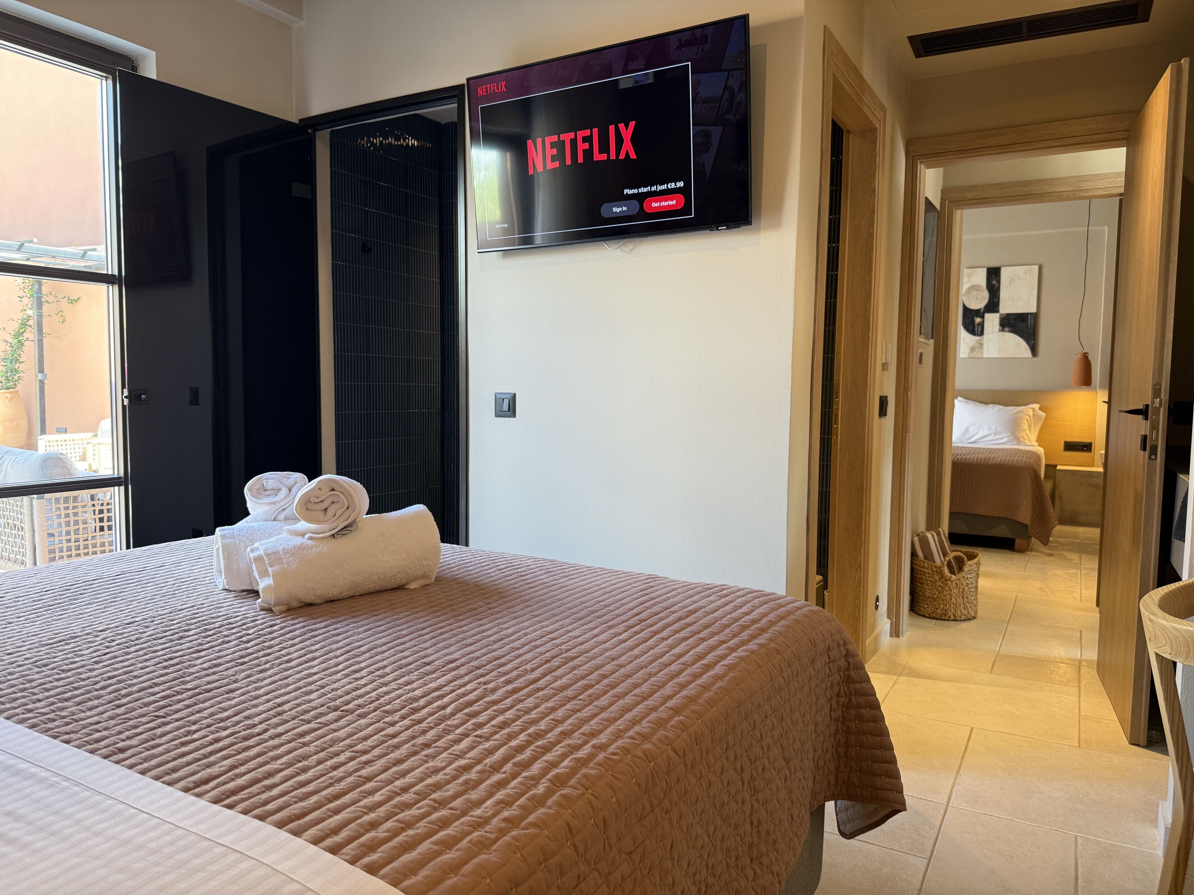 Une chambre d’hôtel avec un lit, un téléviseur fixé au mur diffusant Netflix, une fenêtre et une porte donnant sur une autre pièce.