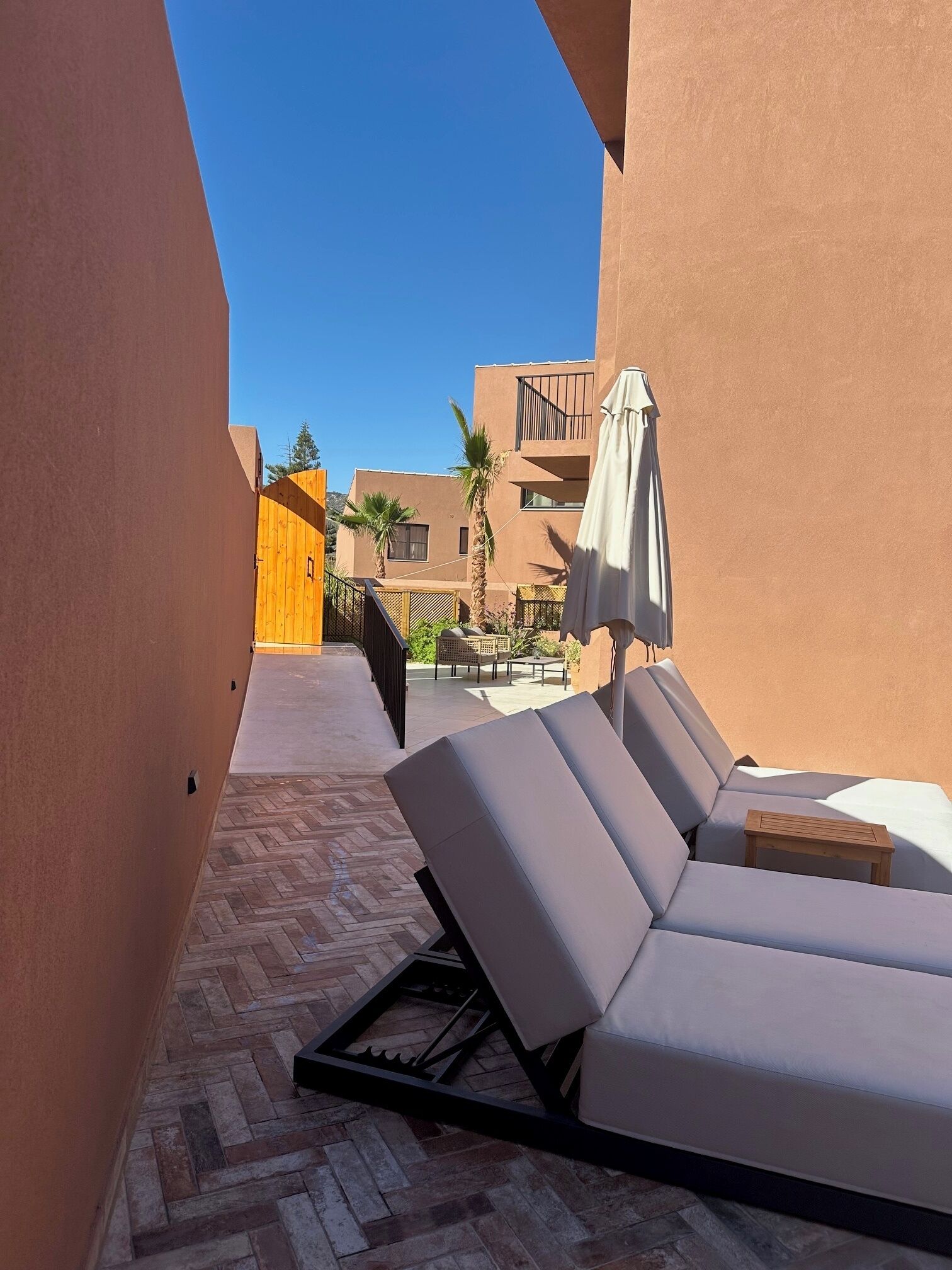 A sunlit outdoor lounge area with a tiled floor, a wooden gate, and a poolside seating area with a table and umbrella.