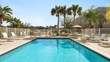 A rectangular swimming pool with clear blue water, surrounded by white lounge chairs and palm trees.