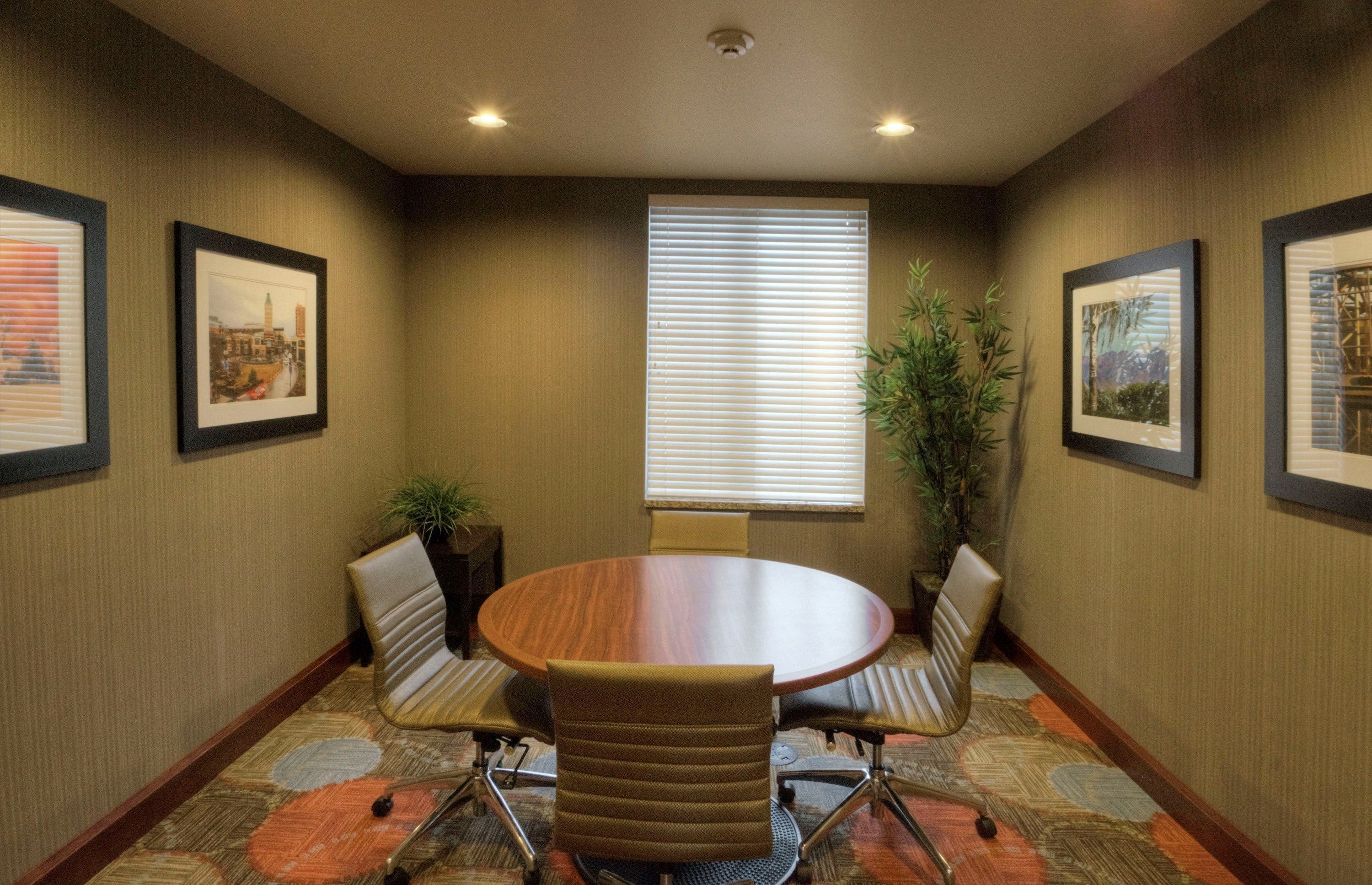Uma sala de conferências com uma mesa redonda, quatro cadeiras, quadros nas paredes e uma janela com persianas.
