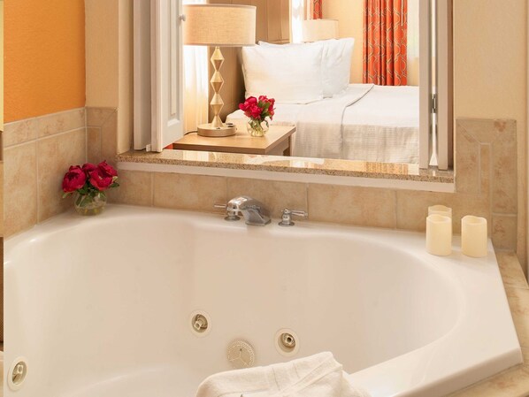 A hotel bathroom with a large bathtub, a mirror reflecting a bed with white bedding, and a vase with red flowers on the counter.